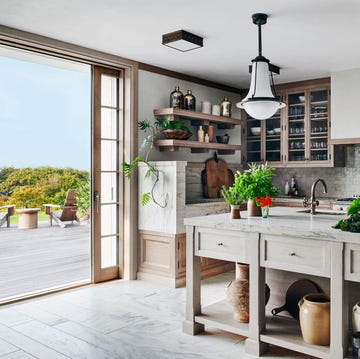 kitchen with an open door to a terrace, large marble topped island with fluted edge, sink, vases stored underneath, two pendants, long countertop with wood cupboards below and glass cabinets above
