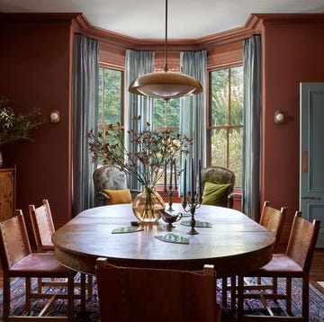 dining room with dark terra cotta walls and light blue gray curtains and paint trim, oblong wood table with chairs with leather seats, pendant above, fireplace with ornate surround, carved wood sideboard, patterned rug