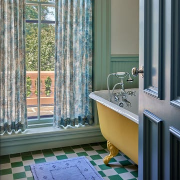 soft white bathroom with sage trimp and wainscoting, claw foot tub painted mustard yellow, silver fittings, large window with translucent floral curtains, green and white checked floor, blue runner, blue gray door