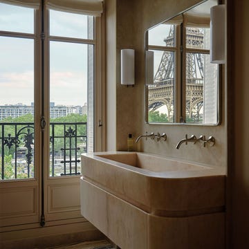 a bathroom has a rectangular light marble sink with curved edges, two faucets, and two sconces flanking a mirror that reflects the eiffel tower, windowed doors open to a small balcony, patternd rug
