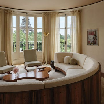 a curved living room has a large circular wood frame sofa with light colored cushions and ball shaped accent pillows, an amorphous wood cocktail table, two armless chairs with cushions, windows with a view of eiffel tower