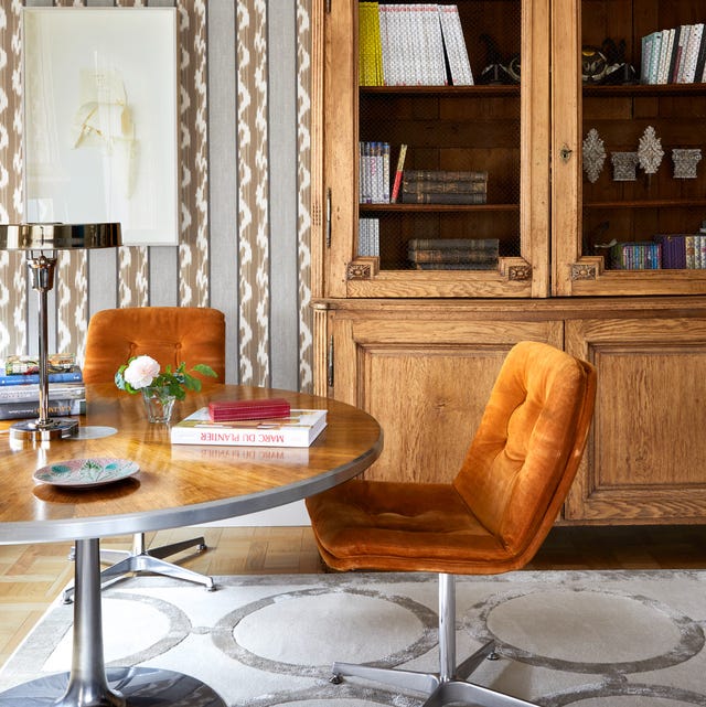 A-List 2024 - ELLE DECOR seating area with round pedestal table and two orange velvet armless chairs on swivels and a large oak cabinet with glass doors behind it and a white on white rug with round designs on the floor