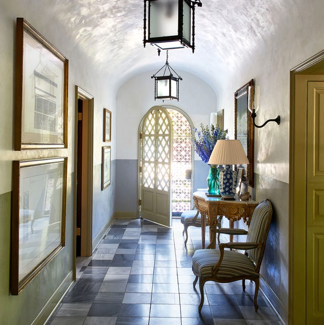 A-List 2024 - ELLE DECOR entry hall with variegated gray tile and artwork on the walls and a gilded demilune table with armchairs flanking it and an oval arched door with screen at the far end and lantern hanging overhead