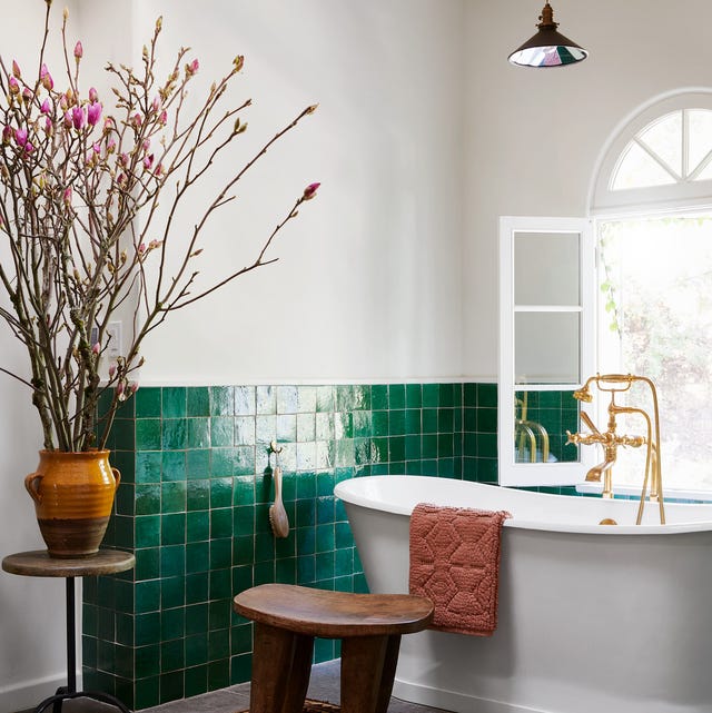 A-List 2024 - ELLE DECOR bathroom with half green shiny tile and standalone tub with gold faucets and a wood low stool next to it and a red patterened rug and a window at back with a view