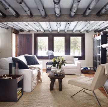 cozy living room with rustic white pine ceiling beams and modern furniture and a stone fireplace and a three panel window at the back