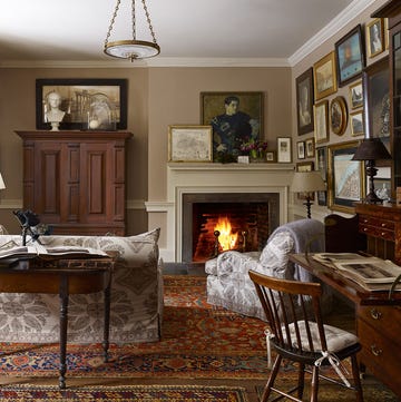 cozy room with several colorful rugs and a lit fireplace at the far end with paintings and framed prints around the wall and a windsor chair at a desk and an armoire and a sofa with a demilune table behind it