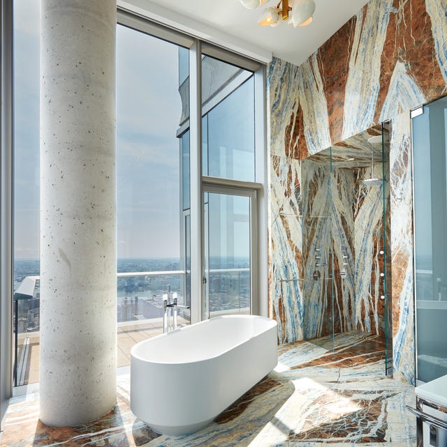 A-List 2024 - ELLE DECOR a standalone tub in a bathroom covered in floor to ceiling blue and white and brown marble with a white column and a glass door and window that look out over a very high and far city view