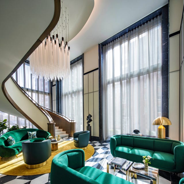 A-List 2024 - ELLE DECOR dramatic double height curtained living area with a sensuous curved staircase leading upstairs and a black and white tile floor and green curvy sofas and a yellow carpet