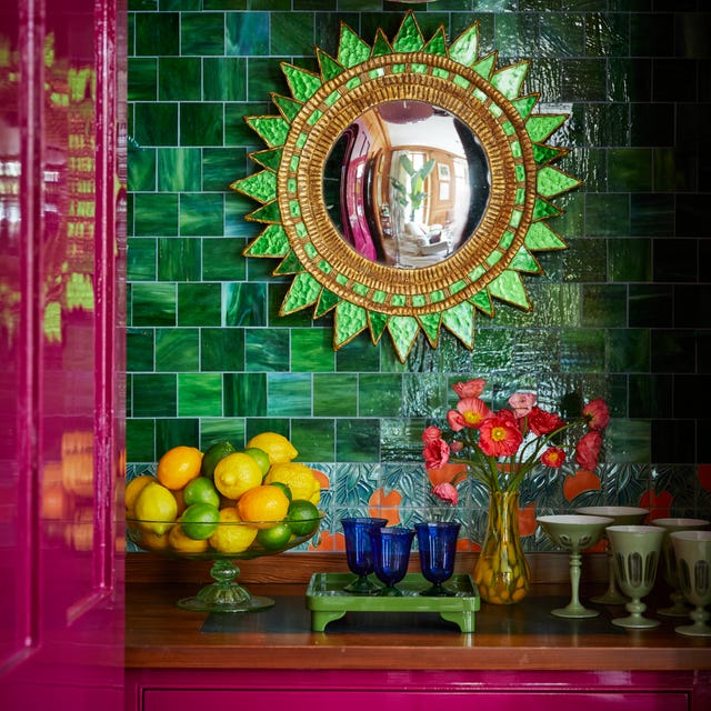 a green sunflower mirror on a green tiled wall in a cubby with bright eggplant magenta laquered drawers and doors and a counter with a bowl of lemons and limes and blue glasses and a yellow vase a green sunflower mirror on a green tiled wall in a cubby with bright eggplant magenta laquered drawers and doors and a counter with a bowl of lemons and limes and blue glasses and a yellow vase