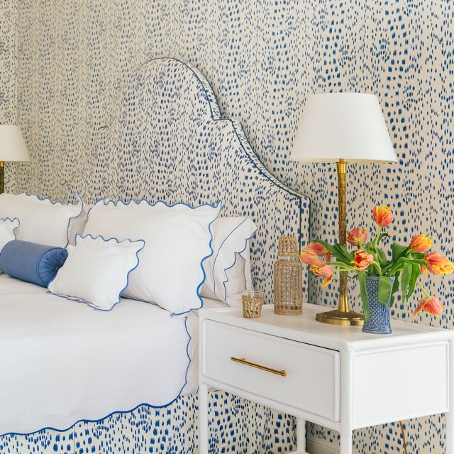 A-List 2024 - ELLE DECOR a bed with a white blue speckled bed headboard and matching wall and bed skirt and white linens edged in matching blue and a small side table with flowers and a lamp