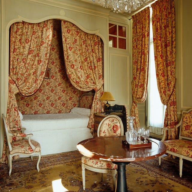 A-List 2024 - ELLE DECOR an inset captains bed with red patterned wallpaper behind it and matching curtains and draperies and upholstery on the chairs and sette and a round pedestal table somewhat at centere
