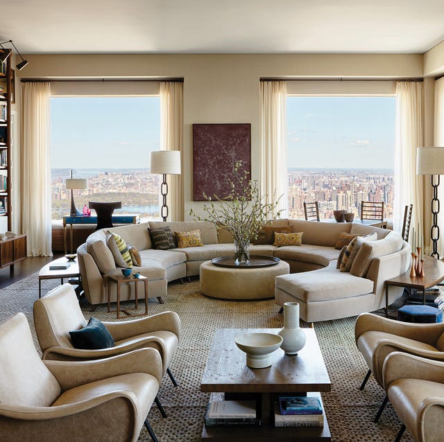 A-List 2024 - ELLE DECOR very off white and white living room with a nearly circular curvy sofa with round cocktail table ottoman at center and numerous chairs and tables and large high windows at back with a view of the city