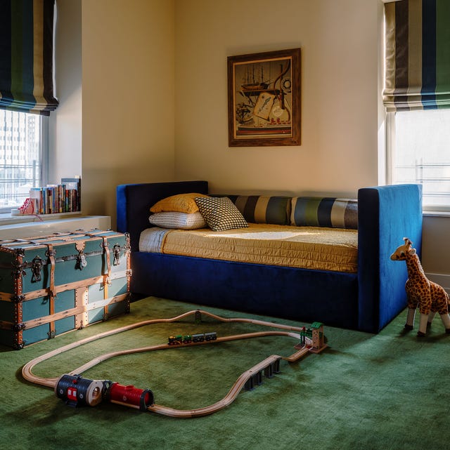 A-LIst 2024 - ELLE DECOR play room with blue velvet daybed with yellow cover and striped bolsters and an old blue strappy trunk to the left on a green carpet with a wooden train set and a small toy giraffe on the floor next to the daybed just hanging out