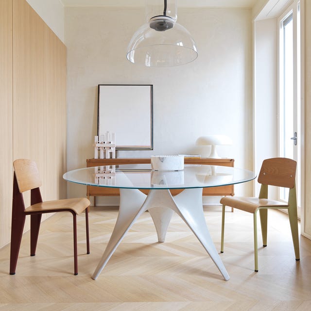 A-List 2024 - ELLE DECOR a stark room with light chevron pattern wood floors and a modern round glass top tripod table with wooden chairs around
